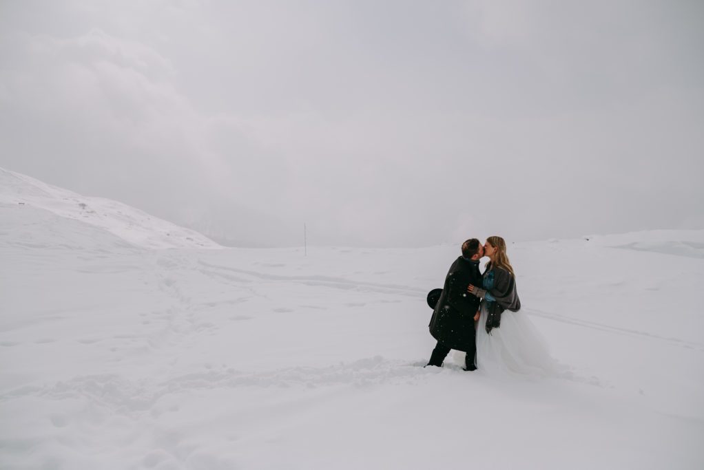 Braut mit Jeansjacke und Bräutigam mit Hut küssen sich im Schneesturm in den Dolomiten