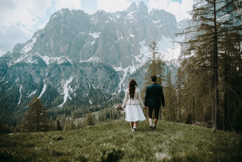 Braut mit Blumenkranz und Bräutigam mit Tiroler Tracht gehen Händchen haltend die Wiese hinab zu einem schneebedeckten Berg in den Dolomiten
