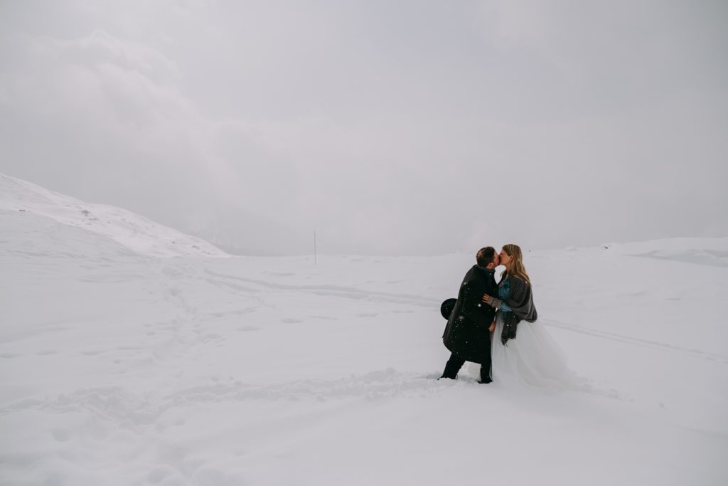 Bride with jeans jacket and groom with hat kissing in the snowstorm in the Dolomites