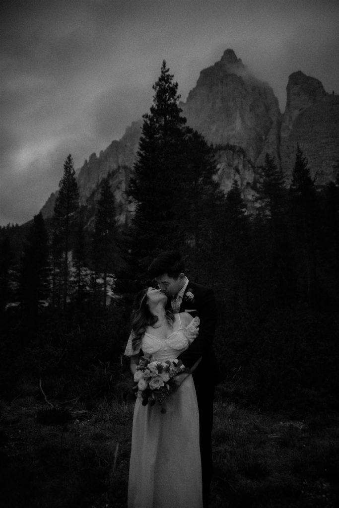 Custom Dolomites Elopement Packages - Black and white photograph of couple kissing in front of a forest and Dolomites mountains in the background